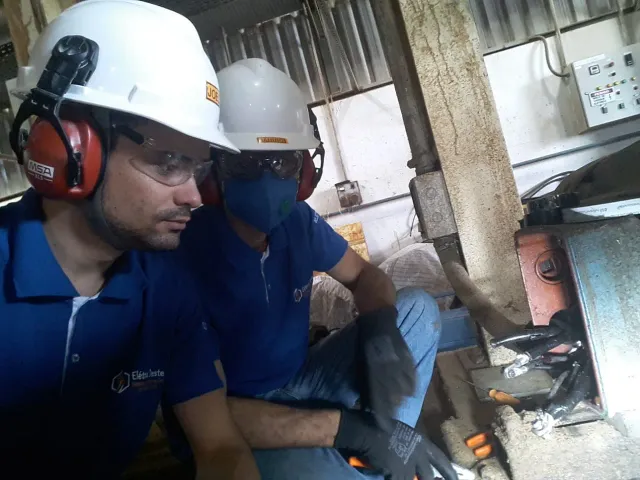 Dois eletricistas da Elétrica Oeste, de capacete e óculos de proteção, agachados junto à caixa de ligação de um equipamento em galpão industrial.
