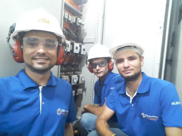 Equipe da Elétrica Oeste uniformizada em sala elétrica, com o painel de comando aberto ao fundo.